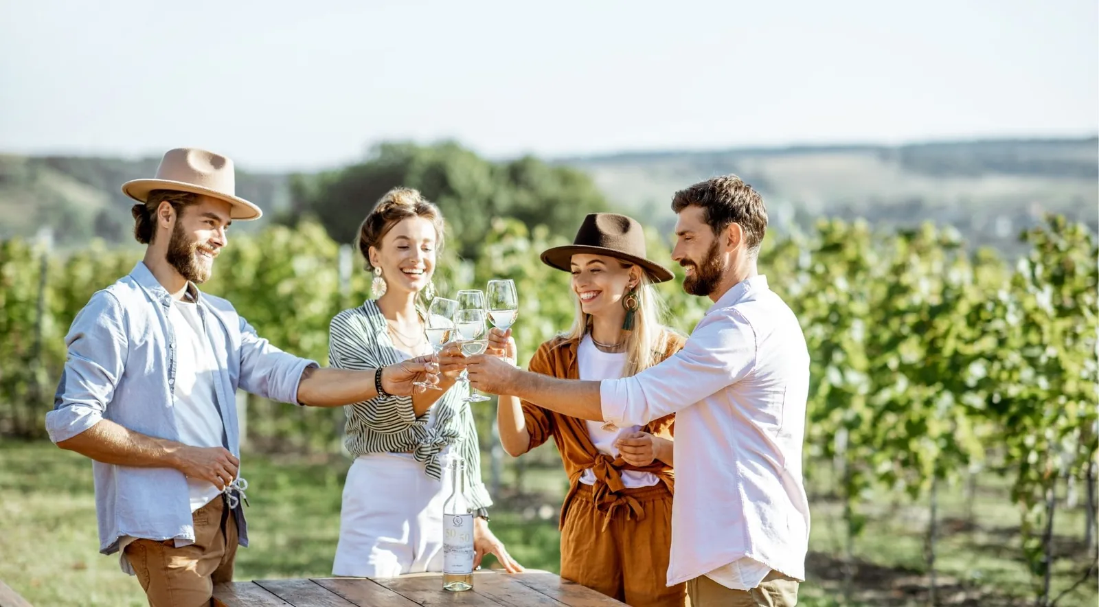 Four friends in sun hats raising glasses of white wine around a table set among vine rows