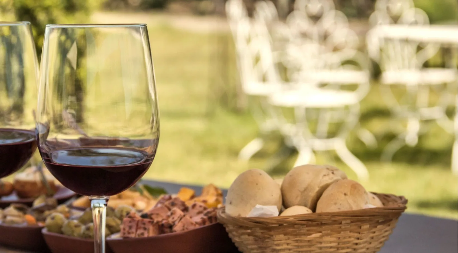 A glass of red wine and a basket of bread rolls on a table set outdoors beside a lawn