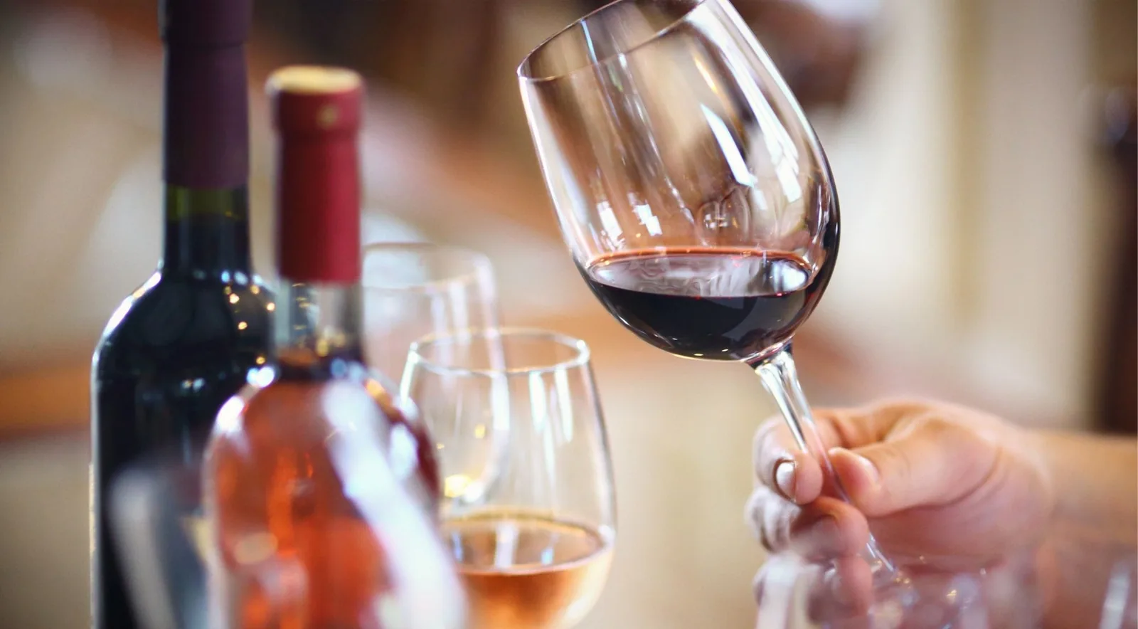 A hand tilting a glass of red wine beside open bottles of red and rose on a tasting table