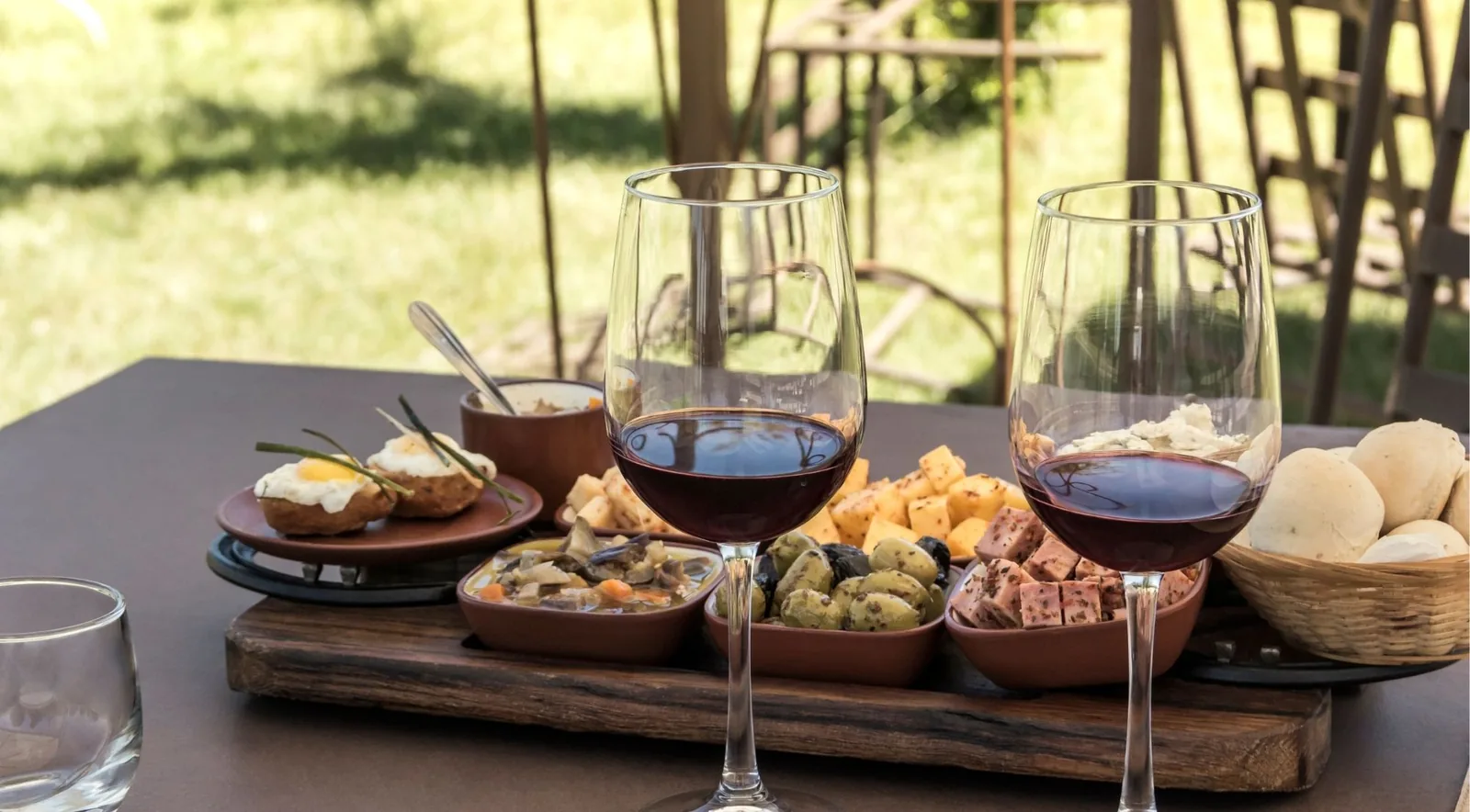 Two glasses of red wine beside a wooden board of cheeses, olives, cured meats and bread